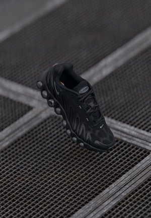 Czarny sportowy sneaker z detalami podeszwy z pęcherzykami, sznurówkami i logo na tle metalowej siatki.