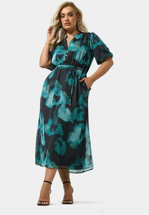 Kromme vrouw met blond haar draagt een turquoise en zwarte bloemen midi-jurk met pofmouwen en zwarte enkellaarzen met bandje, poseert tegen een effen achtergrond.