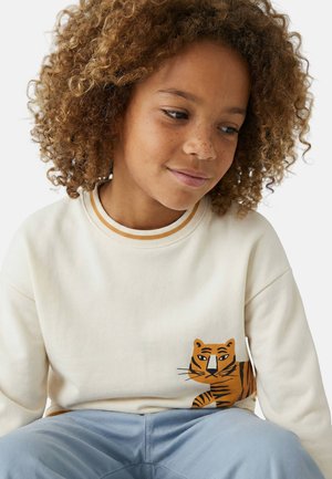 Enfant aux cheveux bouclés portant un sweat-shirt crème clair avec un tigre orange illustré sur le côté, assorti à un pantalon bleu clair.