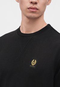 Νέος άνδρας που φοράει μαύρο φούτερ Belstaff με ένα μικρό κίτρινο και μαύρο λογότυπο στο στήθος, εμφανιζόμενος από το λαιμό έως το πάνω μέρος του κορμού.