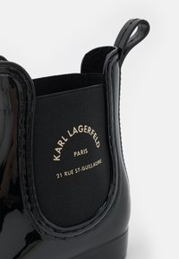 Botín de charol negro con panel lateral elástico y tirador, con el texto "Karl Lagerfeld Paris 21 Rue St-Guillaume" en dorado.