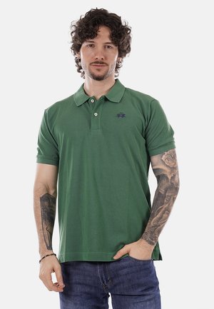 Uomo con capelli ricci che indossa una polo verde e jeans blu, che mostra tatuaggi dettagliati su entrambi gli avambracci.