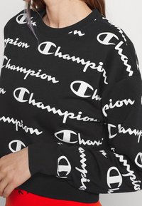 Czarna bluza z białym logo "Champion" w różnych rozmiarach, z okrągłym dekoltem i ściągaczami przy mankietach. Wykonana z miękkiego materiału bawełnianego.