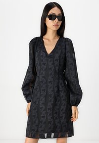 Robe noire avec un décolleté en V, des manches bouffantes et un motif texturé de lignes ondulées. Détails en tissu transparent et silhouette ajustée. Lunettes de soleil noires.