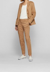 Kobieta w garniturze w kolorze karmelowym z dopasowaną marynarką i spodniami, białej, prążkowanej bluzce oraz białych, niskich trampkach, stojąca na neutralnym tle.