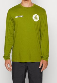 Maglietta verde a maniche lunghe con scollo rotondo, caratterizzata da un logo circolare bianco e un'etichetta nera "BURTON" sul lato sinistro. Tessuto morbido.