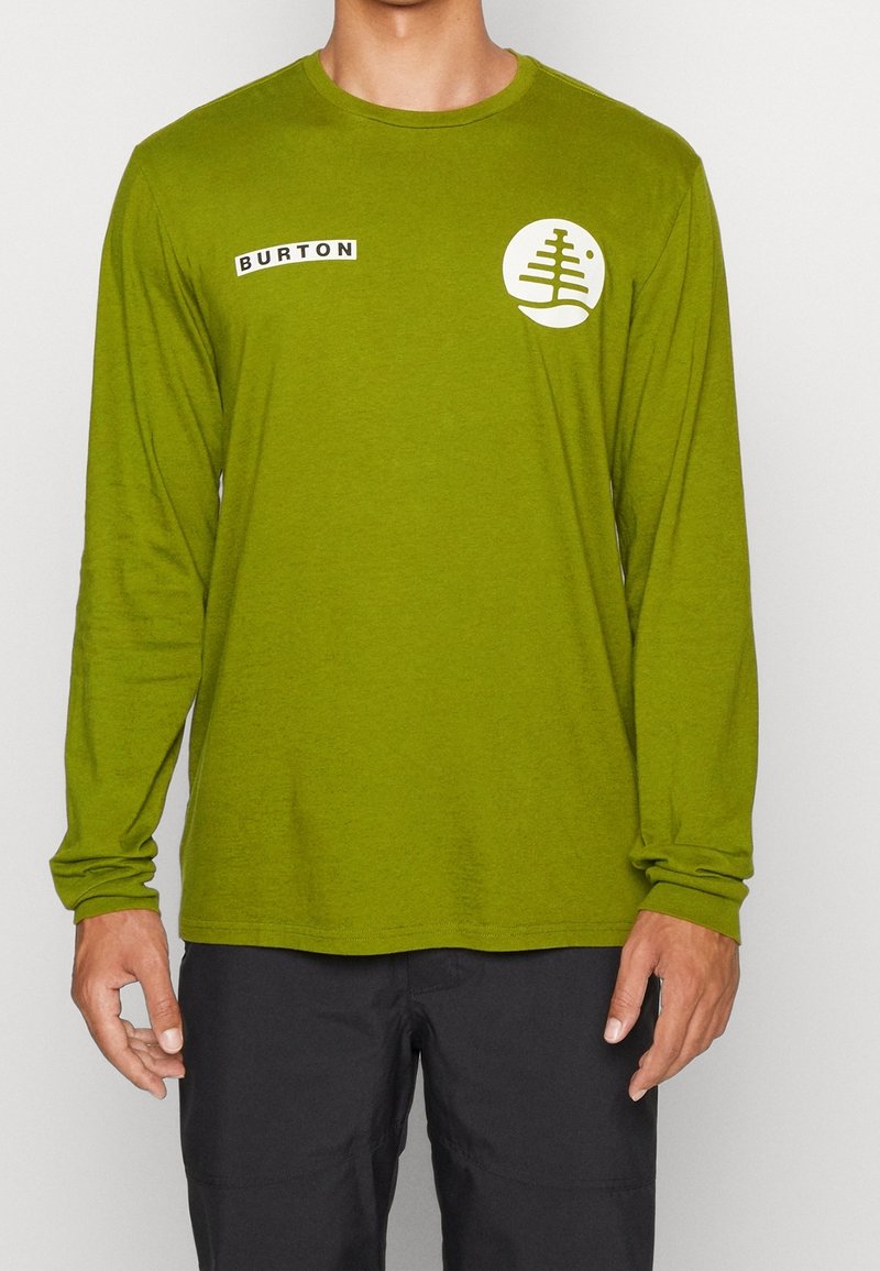 Maglietta verde a maniche lunghe con scollo rotondo, caratterizzata da un logo circolare bianco e un'etichetta nera "BURTON" sul lato sinistro. Tessuto morbido.