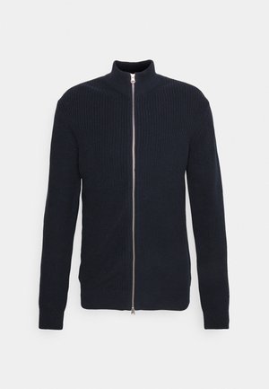 Sininen zip-up kardigan, jossa on ribbitekstuuri, korkea kaulus ja pitkät hihat. Keskellä on hopeinen vetoketju ja tiukat hihansuut.