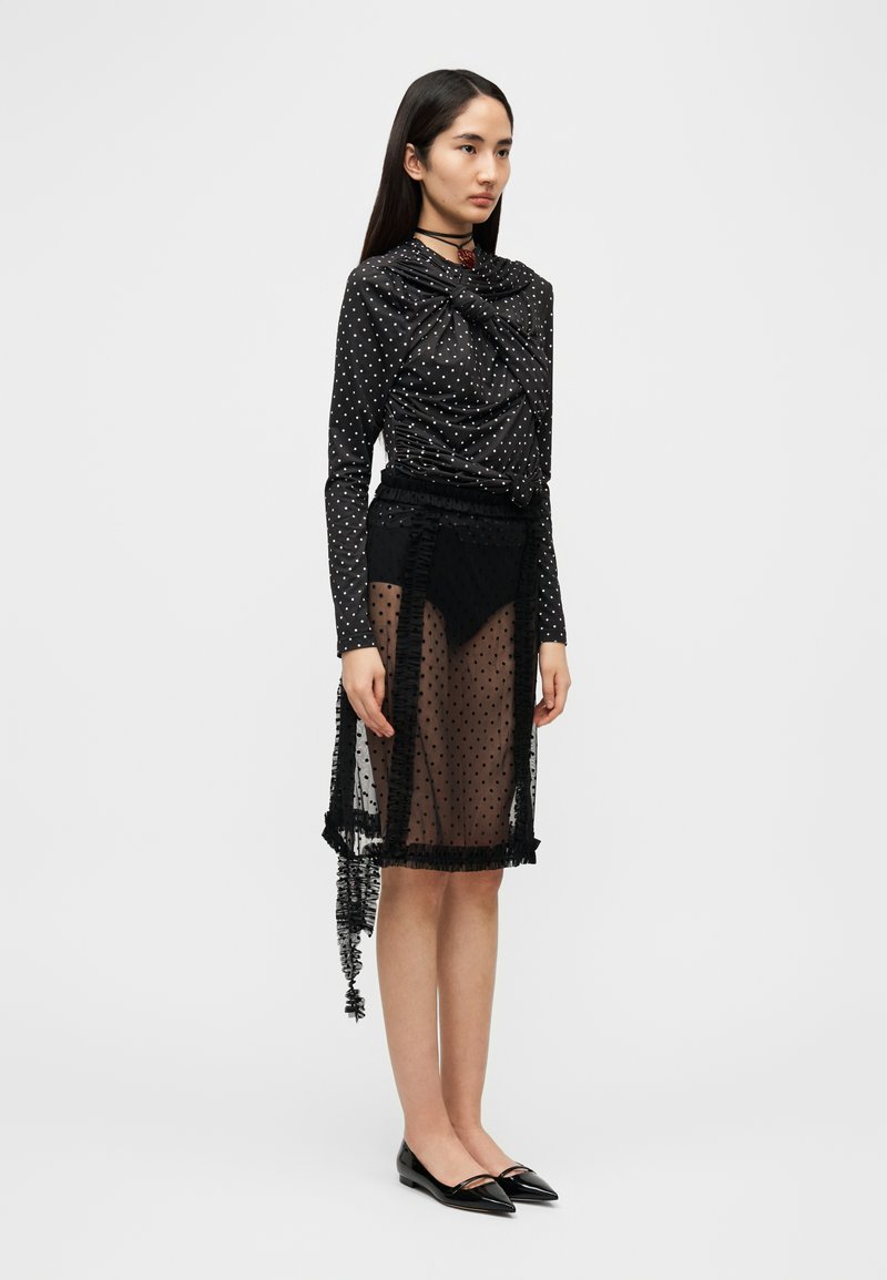 Blouse noire à pois avec un design drapé, associée à une jupe noire transparente ornée de bords à volants et d'un ourlet asymétrique. Chaussures plates noires.