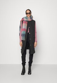 Colete longo acolchoado preto com botões dourados, combinado com um lenço xadrez multicolorido. Modelo veste calças pretas e botas de salto.