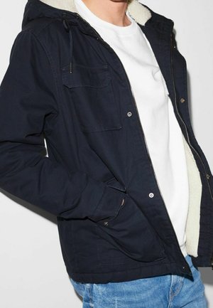 Homme portant une veste à capuche bleu marine non zippée avec une doublure en polaire, un t-shirt blanc à col rond et un jean bleu clair, main dans la poche.
