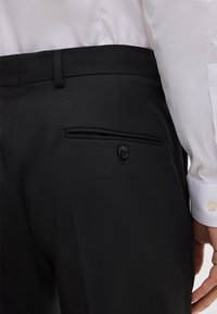 Nahaufnahme einer Person, die schwarze maßgeschneiderte Hose mit einer geknöpften Gesäßtasche und einem in die Hose gesteckten weißen Hemd trägt.