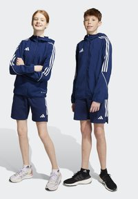 Donkerblauwe sportjack met witte strepen, gecombineerd met bijpassende shorts. Beide dragen sneakers; de ene is grijs, de andere is zwart.