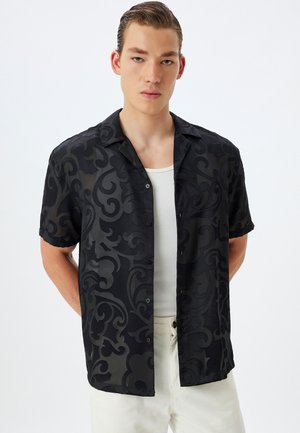 Camisa negra de manga corta con un patrón de remolino tonal, con botones al frente y un corte relajado, usada sobre una camiseta blanca sin mangas, combinada con pantalones de colores claros.