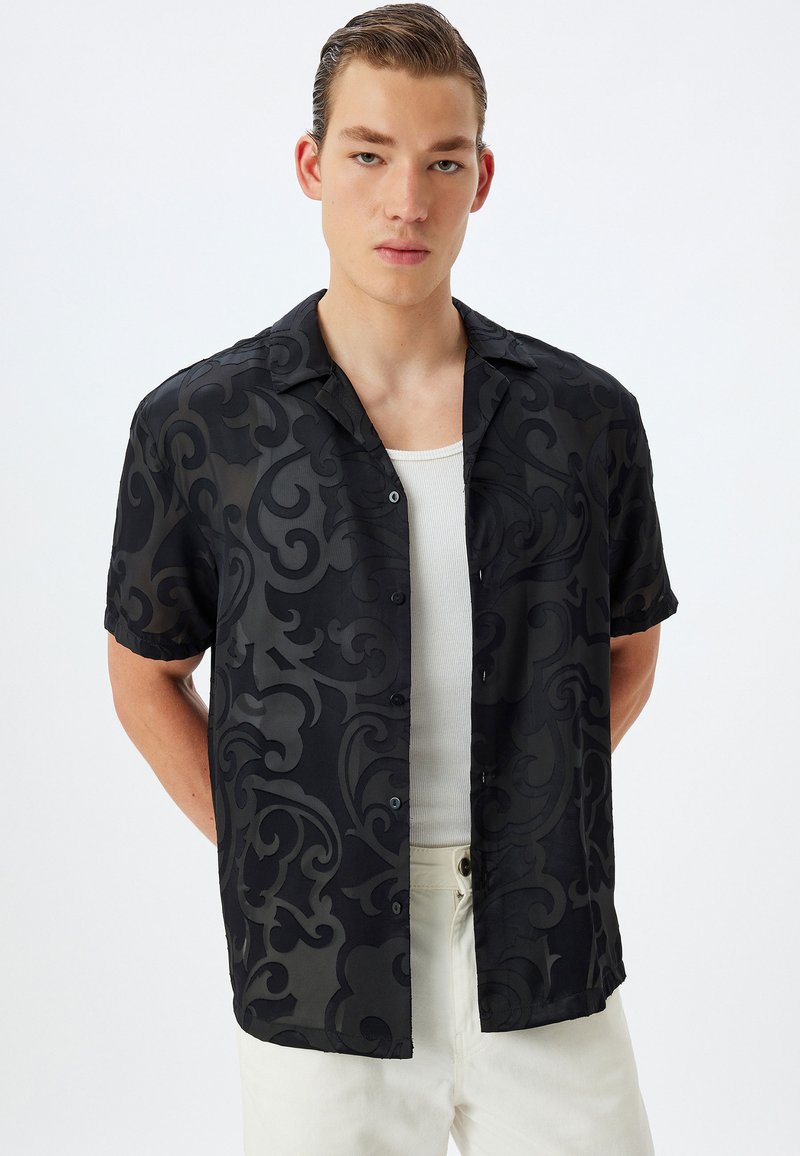 Camisa negra de manga corta con un patrón de remolino tonal, con botones al frente y un corte relajado, usada sobre una camiseta blanca sin mangas, combinada con pantalones de colores claros.