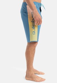 Modré plavky s gradientovým bočným panelom v žltej a ružovej farbe, s veľkým čiernym logom "Quiksilver" a nastaviteľnou šnúrkou.