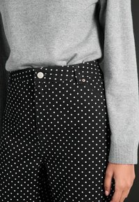 Jeans negre cu buline albe au o croială slim, talie din material și detalii cu nasture argintiu. Deasupra este purtat un pulover gri cu mansete cu nervuri.