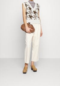 Dzianinowa kamizelka na drutach z dekoltem w serek, zdobiona brązowo-kremowymi wzorami geometrycznymi. W zestawie kremowe spodnie zwężane i brązowe chodaki; trzyma miękką kopertówkę.