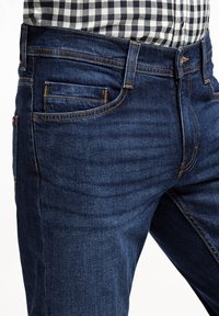 Granatowe dżinsy z tradycyjnym pięciopocketowym fasonem, widocznym szwem w kontrastującym żółtym kolorze i dopasowanym kroju.