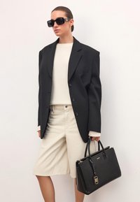 Fekete, túlméretezett blazer, krém színű pulóver, bézs sort, nagy fekete táska texturált felülettel és arany kiegészítőkkel, valamint sötét, szögletes napszemüveg.