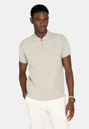 Joven con cabello corto, vestido con una camiseta tipo polo beige claro con cremallera y pantalones blancos, de pie con una mano en el bolsillo.