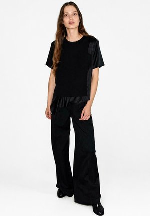 Joven mujer con cabello largo y castaño, vestida con una camiseta negra de manga corta y pantalones negros de pierna ancha, de pie contra un fondo blanco.