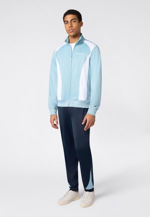 Man draagt een lichtblauw-wit jack met rits, donkerblauwe trainingsbroek met lichtblauwe accenten, en witte sneakers, staand tegen een egale achtergrond.