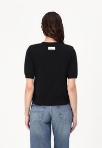 Schwarzer kurzärmeliger Pullover mit rundem Ausschnitt und einem Etikett auf der Rückseite. Glatte Oberfläche; taillierte Passform kontrastiert mit lockeren Jeans.