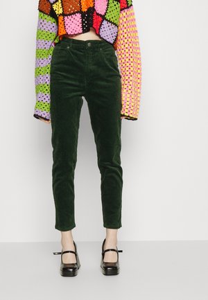 Person wearing dark green high-waisted corduroy pants, colorful patchwork crochet sweater, and black Mary Jane platform heels standing against a white background.