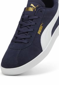 Tengerészkék velúr edzőcipő fehér részletekkel, textúrált felülettel, lapos fűzőkkel és arany "Puma" logóval az oldalon.