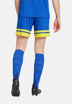 Blaue Sportshorts mit gelben Akzenten, kombiniert mit blauen knielangen Socken und schwarzen Fußballschuhen mit goldenen Details.