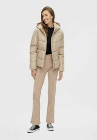 Beige pufferjakke med hette, med quiltet tekstur og elastiske mansjetter, båret over en svart topp og parret med beige bukser og svarte joggesko.