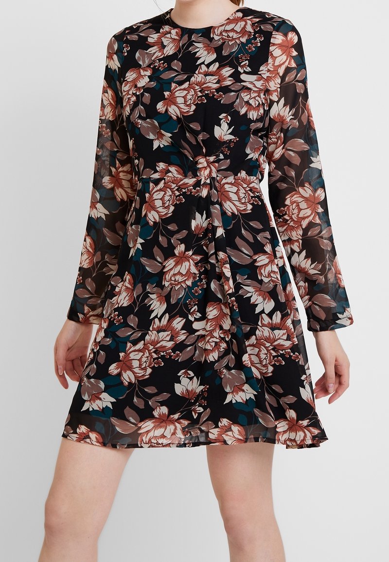 Robe longue à manches longues noire à fleurs roses, beiges et bleu sarcelle, taille froncée et manches transparentes, portée à mi-cuisse.