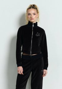 Blondynka stoi przy ścianie, ubrana w czarną kurtkę na zamek i pasujące spodnie z cyrkoniowymi logo "LH", włosy w wysokim kucyku.