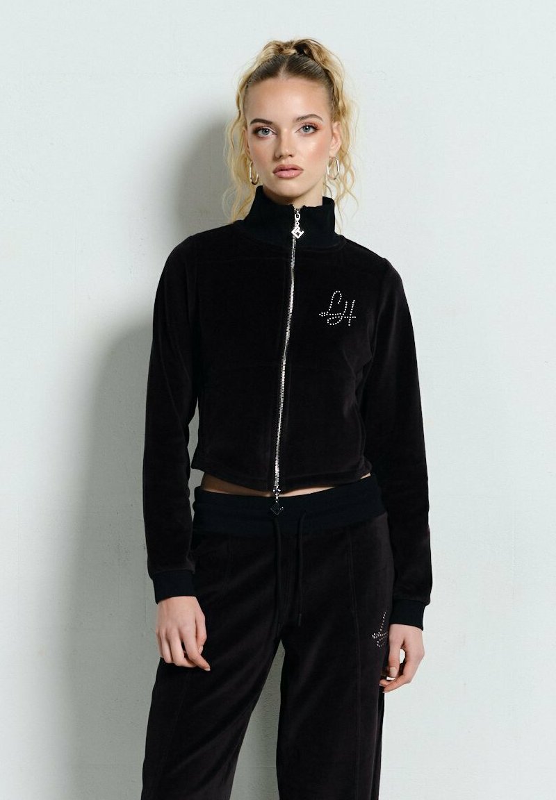 Blondynka stoi przy ścianie, ubrana w czarną kurtkę na zamek i pasujące spodnie z cyrkoniowymi logo "LH", włosy w wysokim kucyku.