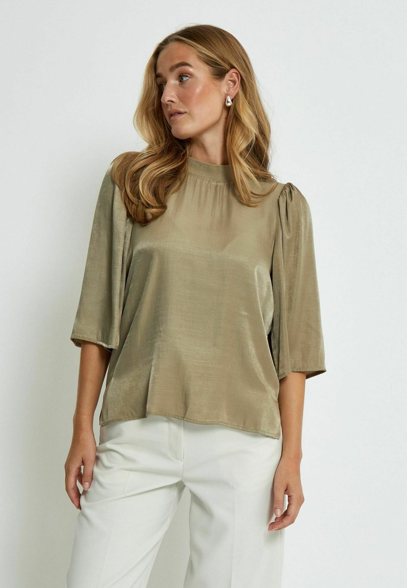Olive-green satin blouse with a high neckline, puffed sleeves, and a relaxed fit. Smooth texture, softly draping, paired with white trousers.