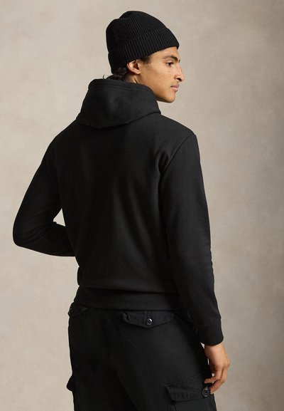 Egy férfi fekete sapkában, fekete kapucnis pulóverben és fekete terepnadrágban áll oldalra fordulva egy egyszínű bézs háttér előtt.