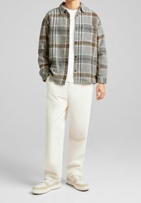 Camisa de cuadros gris y marrón sobre un suéter blanco, combinada con pantalones color crema y zapatillas deportivas beige. Diseño de botonadura con detalle de bolsillo.