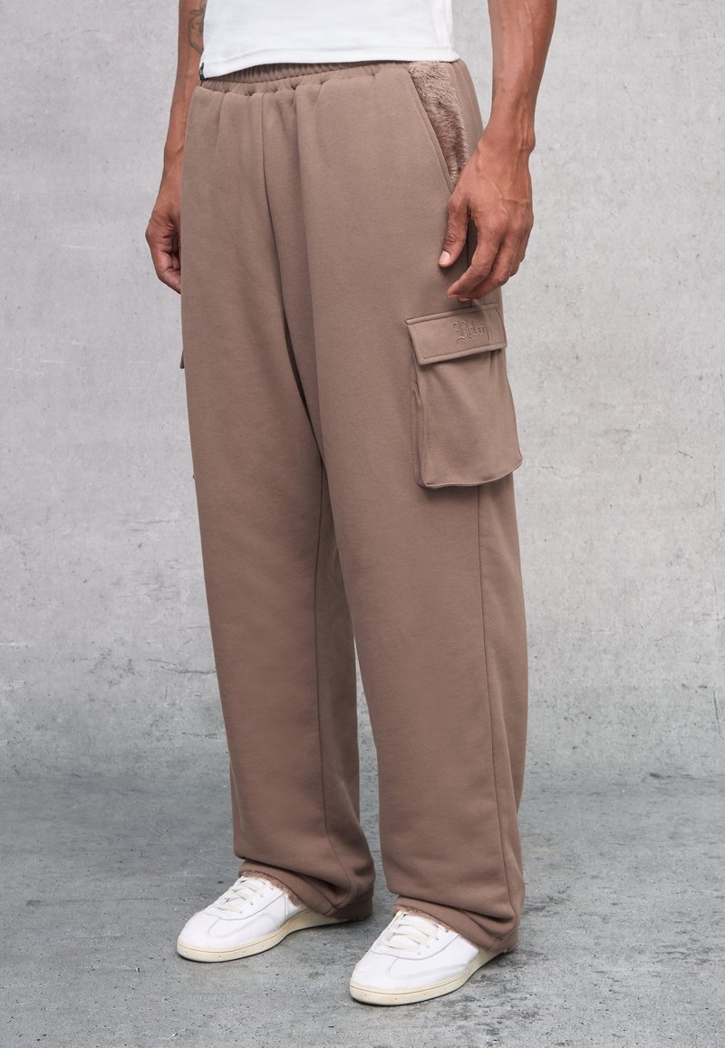 Bruine trainingsbroek met een ontspannen pasvorm, voorzien van cargozakken en een zachte textuur. Gecombineerd met witte sneakers, staand tegen een grijze achtergrond.
