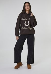 Vrouw in een oversized donker hoodie met een hoefijzer graphic, ruime zwarte spijkerbroek en bruin veterlaarzen, staand tegen een effen achtergrond.