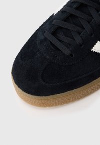Black suede sneaker with textured surface, rubber gum sole, and black laces. White accent stripes on the side.