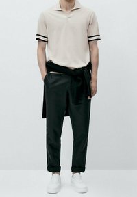 Polo beige avec des accents rayés noirs, associé à un pantalon noir fuselé et des baskets blanches. Le polo est porté avec un pull noir noué à la taille.