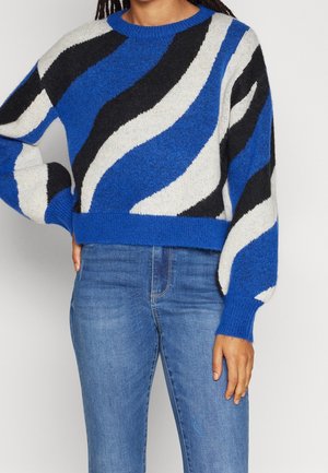 Femme portant un pull à rayures ondulées bleu, blanc et noir à manches longues et un jean bleu, debout avec une main sur la hanche.
