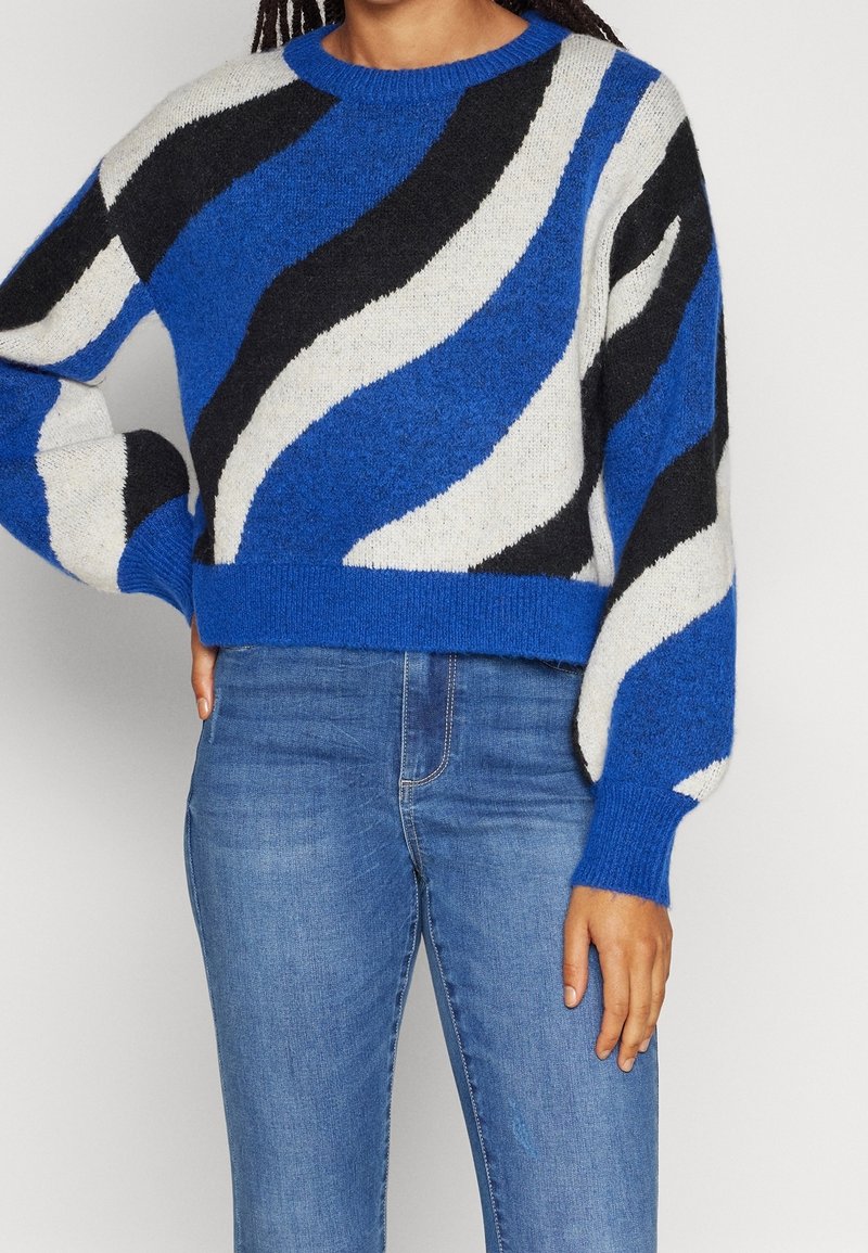 Femme portant un pull à rayures ondulées bleu, blanc et noir à manches longues et un jean bleu, debout avec une main sur la hanche.