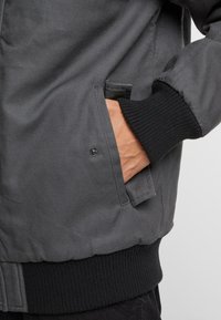 Pilkas bombonešis su juodu gumbu, šonine kišene ir lygia tekstūra. Dėl užtrauktuko uždarymo ir papildomų siūlių detalių.