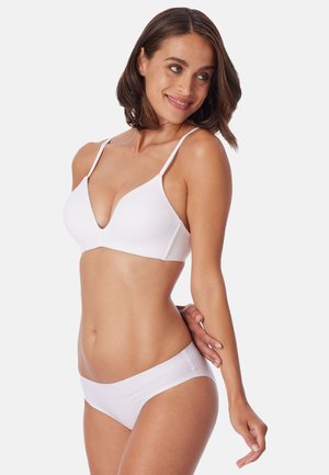 Woman with short brown hair wearing a white wireless bra and matching briefs, smiling and looking to the side against a plain background.