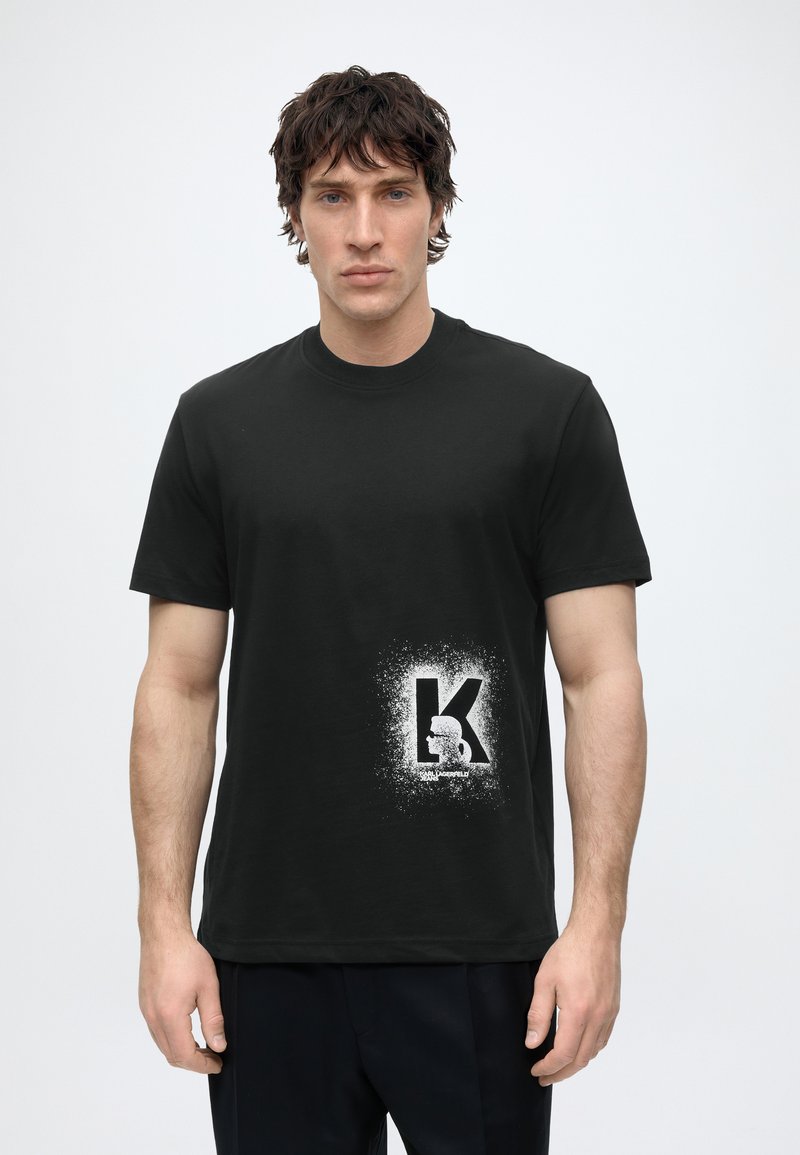 Νέος άνδρας με σκούρα μαλλιά φοράει μαύρο T-shirt με λευκό «K» και αφηρημένο σχέδιο κοντά στο κάτω δεξιό μέρος.