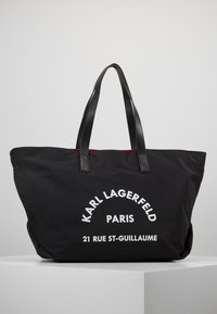 Sac cabas noir avec poignées doubles, texte blanc "Karl Lagerfeld Paris 21 Rue St-Guillaume", présenté sur un piédestal blanc sur un fond uni.