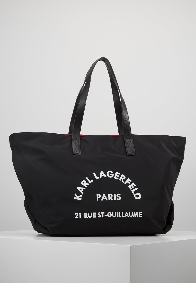 Sac cabas noir avec poignées doubles, texte blanc "Karl Lagerfeld Paris 21 Rue St-Guillaume", présenté sur un piédestal blanc sur un fond uni.