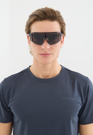 Junger Mann mit dunkler, übergroßer Sportsonnenbrille und einem marineblauen Endurance-T-Shirt, der frontal vor einem einfachen weißen Hintergrund steht.